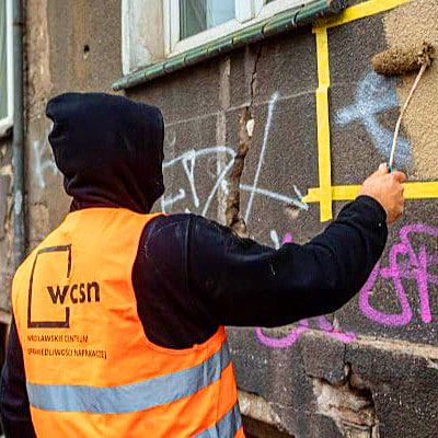 Zdjęcie przedstawiające mężczyznę w bluzie i kamizelce z napisem WCSN malującego wałkiem ścianę budynku